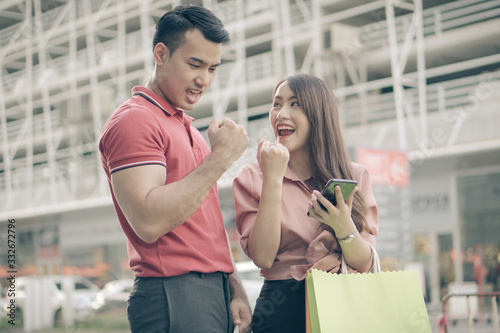 Happy young couple of shoppers walking in the shopping street towards and holding colorful shopping bags in hand and use a smartphone for check promotion. Concept of sale and Black Friday shopping