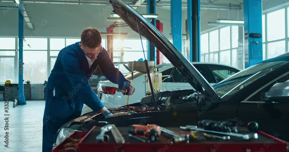 Male Caucasian young auto mechanic standing at open car and repairing motor in big garage. Man in uniform and goggles servicing vehicle in automobile repairment salon indoor. Transport service concept
