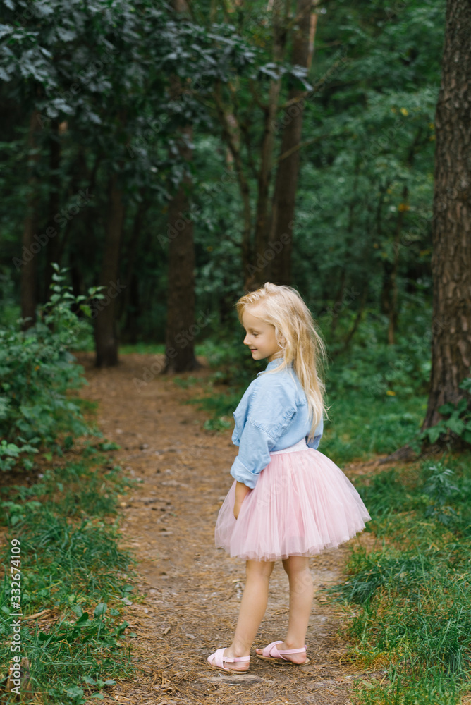 A small blonde girl of five in romantic clothes walks along a forest path and smiles