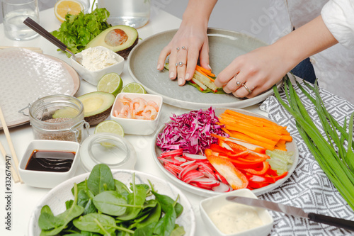 The process of cooking spring rolls - woman's hands putting rolling spring roll with basil, pepper and cucumber. Table with spring rolls ingredients, fresh vegetables, geen onion and avocado. photo