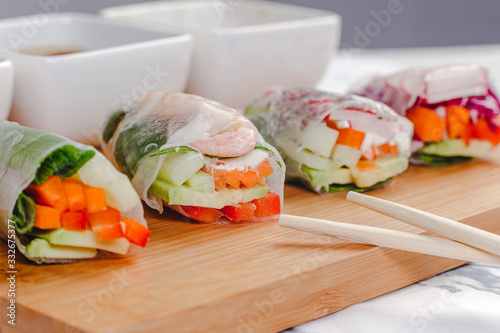 Vertical close up shot of fresh spring rolls with shrimp and vegetables cut in half and different sauces served on a wood plate.