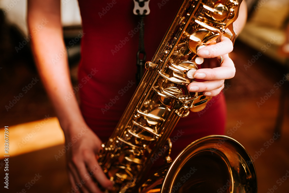 Obraz premium Young beautiful woman smile while playing tenor saxophone in nice cozy condo flat in the sundown with a sythesizer in the background