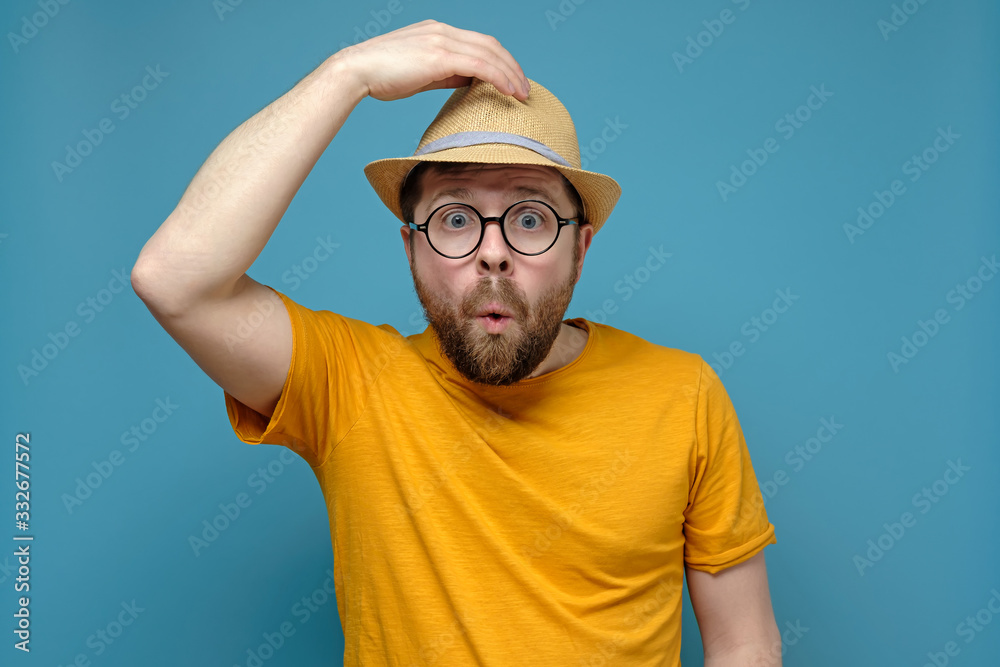 Surprised Caucasian man in round glasses clings to his hat, as if greeting someone, on a blue background.