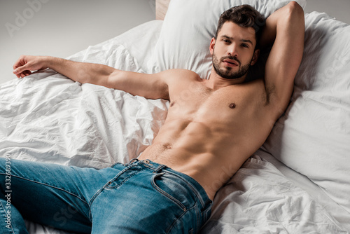 handsome sexy shirtless man in jeans lying on bed on grey