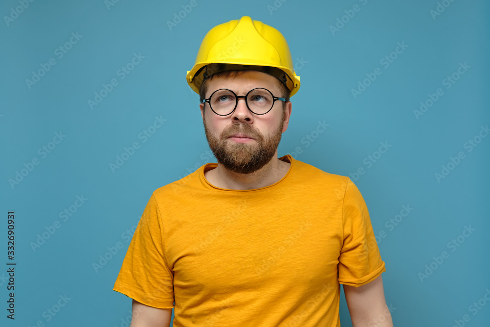 Puzzled bearded man in a construction helmet and round glasses looks up thoughtfully, on a blue background.