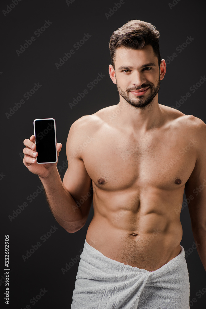 Fototapeta premium smiling sexy man in towel showing smartphone with blank screen isolated on black