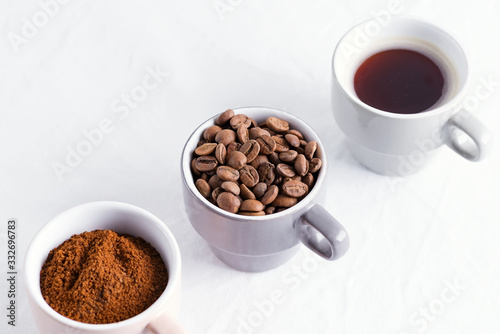 Three cups of different condition of coffee - beans, freshly ground and coffee drink on a marble light grey background.