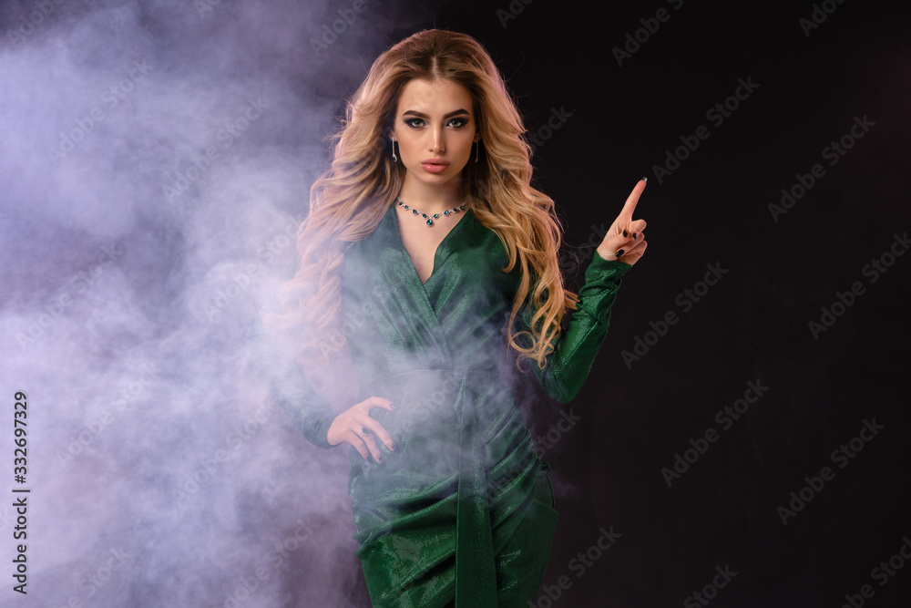 Blonde girl in green stylish dress and jewelry. Pointing at something by forefinger, posing on black smoky background. Close up, copy space