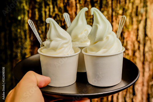 Hand holding three vanilla ice scream paper cups ready to serve on black tray with plastic spoon on wood background. Close up milky soft cream so yummy. photo