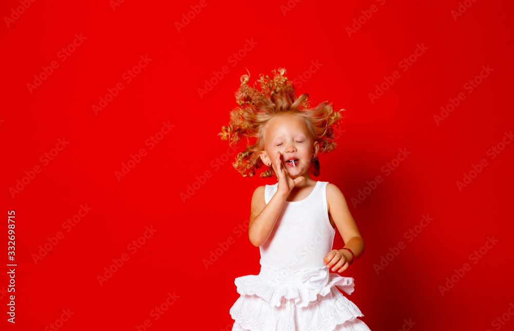 Beautiful little curly blonde girl in a white dress eating lollipop jumping isolated on red