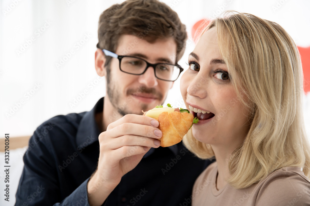 Caucasian young couple portrait in bedroom concept. Boyfriend embracing and feeding his girlfriend a piece of sandwich in bed room in morning. Passionate love of young couple having romantic moment.