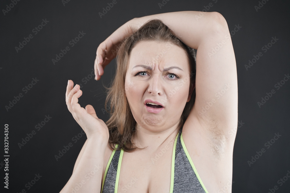 Fototapeta premium unhappy woman shows her unshaved armpit. plus size middle age woman is not happy with hair in her armpits. Caucasian girl is emotional sad and upset. black background in the photo Studio.