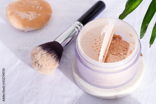 Plastic box with face powder and brush for make-up on a white textile background with green palm leaves.