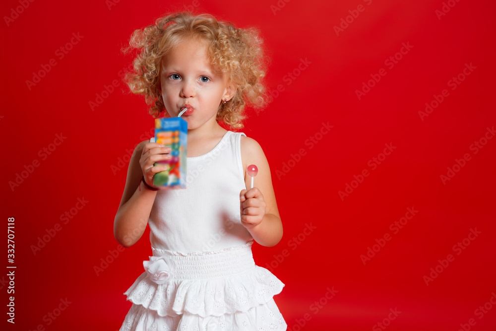 Cute curly girl in white dress drinking juice holding lollipop isolated on red background Cute curly girl in white dress drinking juice holding lollipop isolated on red background