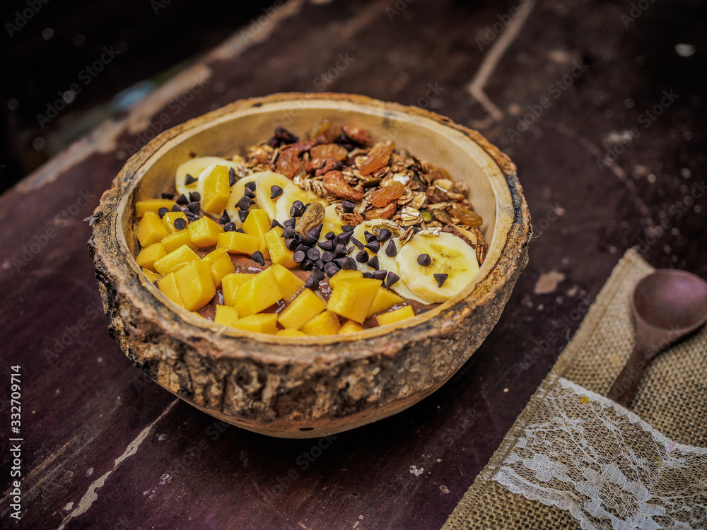 Fototapeta premium close-up of a vegan fruit smoothie bowl with wooden spoon, granola, nuts, banana mango and cocoa on a table for a healthy breakfast 