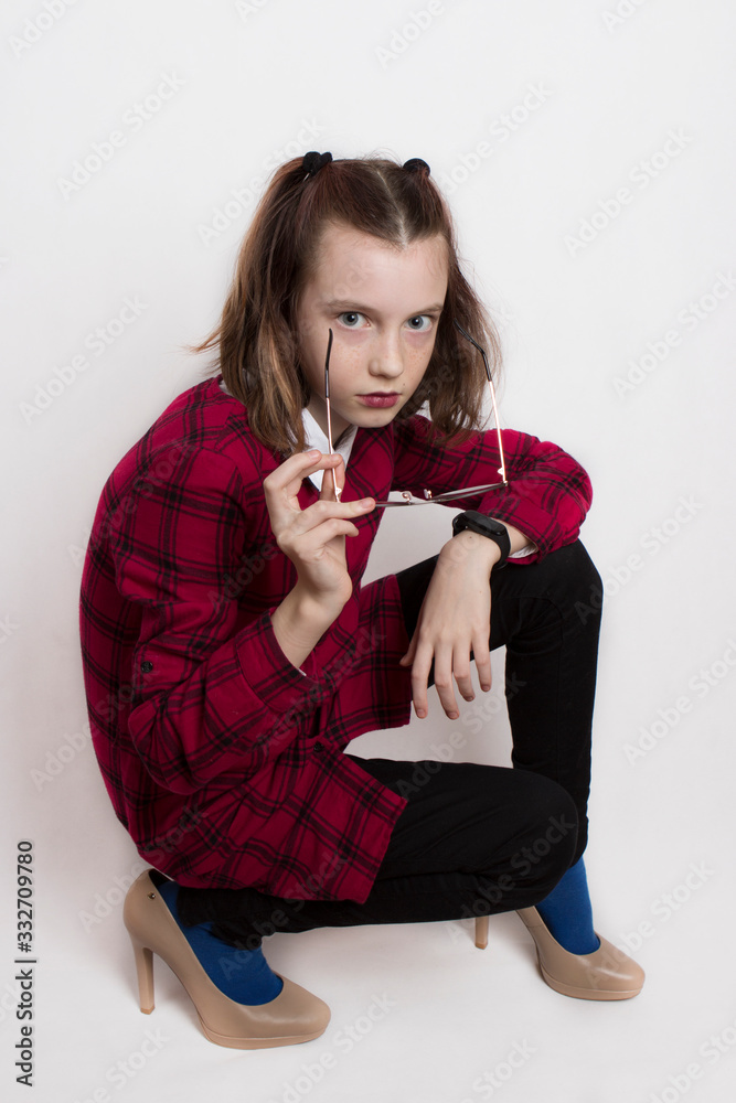 Fototapeta premium a fashionista teenage girl in a red plaid dress dressed mother's high-heeled shoes and glasses and indulges, pretending to be an adult.