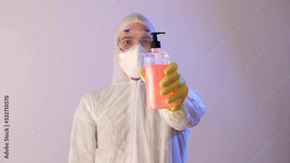Man in a medical mask, protective suit, goggles holds a package with liquid soap or sanitiser gel for clean hands. Wash your hands. Coronavirus or COVID-19. Pandemic worldwide, panic. Man in a medical mask, protective suit, goggles holds a package with liquid soap or sanitiser gel for clean hands. Wash your hands. Coronavirus or COVID-19. Pandemic worldwide, panic.