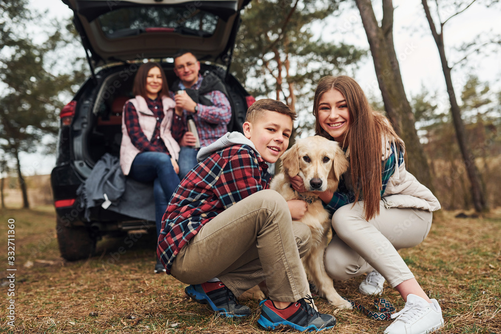 Fototapeta premium Happy family sitting and having fun with their dog near modern car outdoors in forest