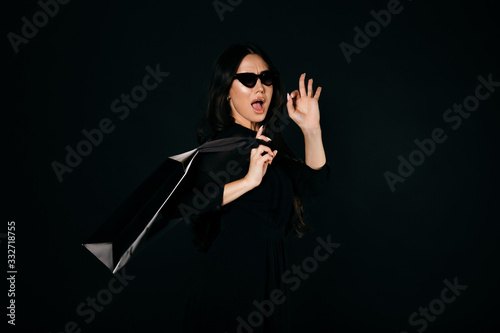 Beautiful girl in sunglasses and black dress with a shopping bag on her back showing ok sign. Isolated on black
