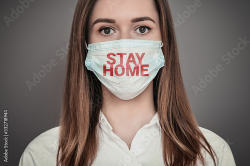 Portrait of a scared young woman wearing a medical respiratory mask with stay home words isolated over dark grey background, coronovirus epidemic, quarantine, pandemic, isolation concept photo