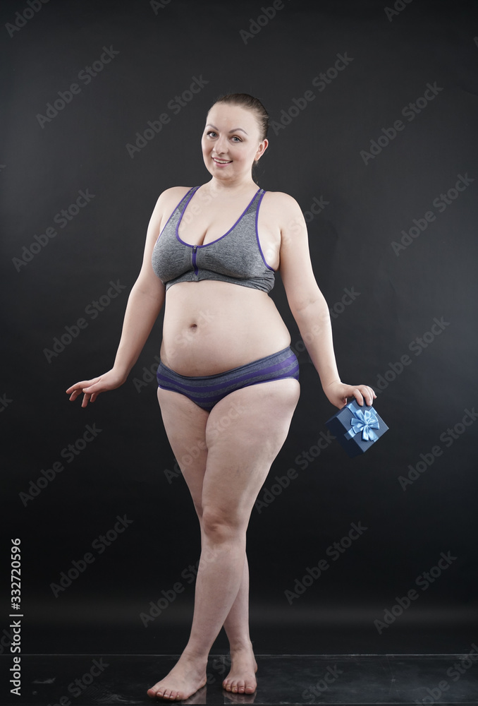 Obraz premium Beautiful plus size woman wearing sport underweaer holds box with gift subscription to a fitness on black studio solid background.