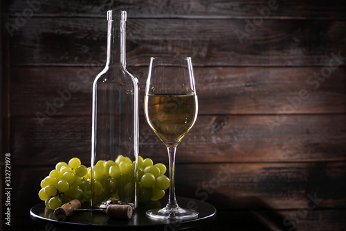 White empty wine bottle and full wineglass with grapes on a table on a wooden background