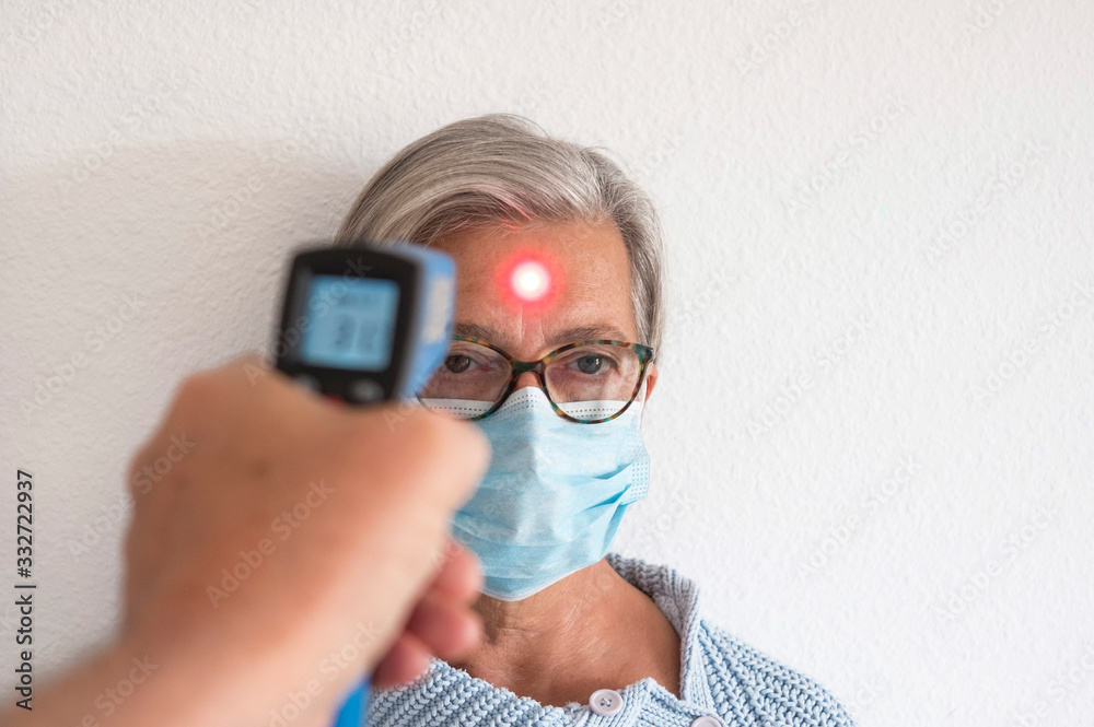 Temperature is checked for a senior woman - a retired person with a mask to avoid COVID-19 coronavirus infection. Prevention and cure concept