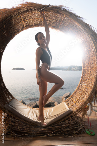 Beautiful girl in black swimsuit posing outdoors on the straw nest over sea and sky background