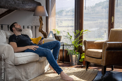 Boring young man sitting on the sofa asleep with his mobile phone in his hand in a rustic house. Home concept