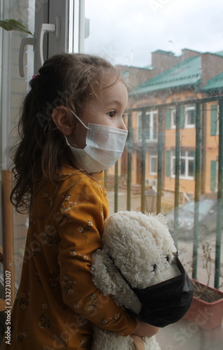 Fotografia A girl and a toy bear in medical masks near the window , looking at the street