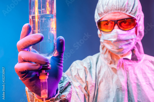 A lab technician in a protective suit , mask, yellow glasses and rubber gloves holds a measuring bottle in his hands.