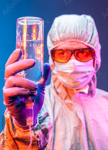 A lab technician in a protective suit , mask, yellow glasses and rubber gloves holds a measuring bottle in his hands.