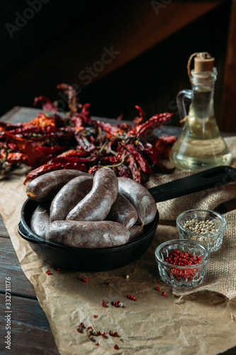 Homemade juicy pork sausages in a black frying pan, spices, pickled tomatoes and dried peppers on a craft paper, on wooden rustic background.