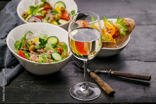 Food still life. White wine in a glass, salads with vegetables and micro-greens in white bowls, canapes with pate and sweet pepper on a dark wooden table.