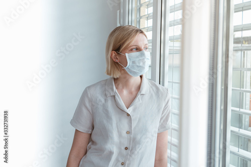 Worried woman looking through the window at home quarantine. Working from home, being alone. Wearing face protection mask in prevention of Coronavirus, ebola, TB and other diseases and viruses outbrea photo