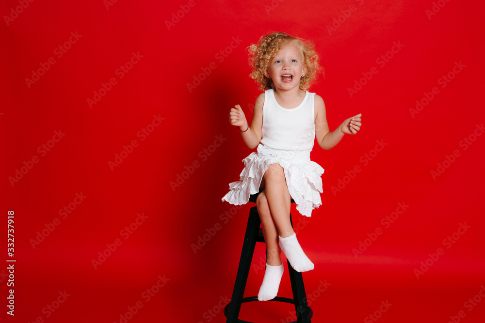 Amuzing funny little blonde girl in white dress sitting on chair isolated on red Amuzing funny little blonde girl in white dress sitting on chair isolated on red