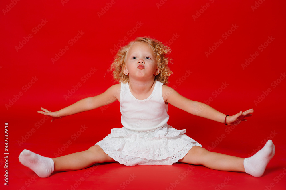 Beautiful little curly blonde girl makes kissing face on the floor isolated on red