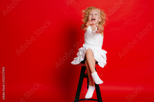 Amuzing funny little blonde girl in white dress sitting on chair isolated on red photo