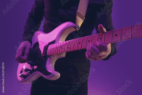 Young and joyful caucasian musician playing guitar on gradient purple studio background in neon light. Concept of music, hobby, festival. Colorful portrait of modern artist. Attented and inspired.