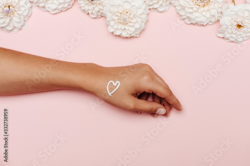 Woman's hand with a white cream heart, on the top white dahlia flowers on pink background. The concept of self love.