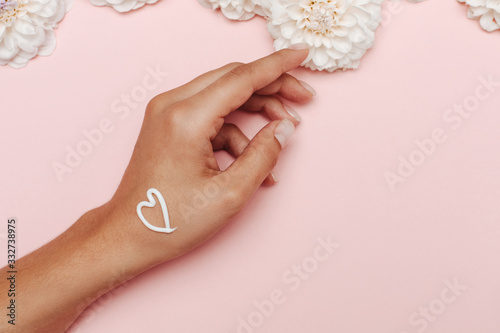 Woman s hand with a white cream heart  on the top white dahlia flowers on pink background. The concept of self love.