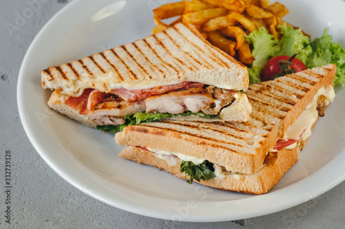 Plate with two chicken and bacon club sandwiches with fries and vegetables on a white plate on a concrete background.