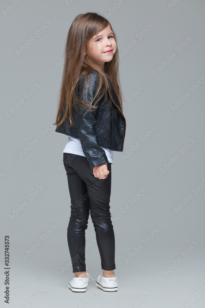 Baby girl with long hair in black leather jacket and leggings is standing in the studio on grey background Baby girl with long hair in black leather jacket and leggings is standing in the studio on grey background