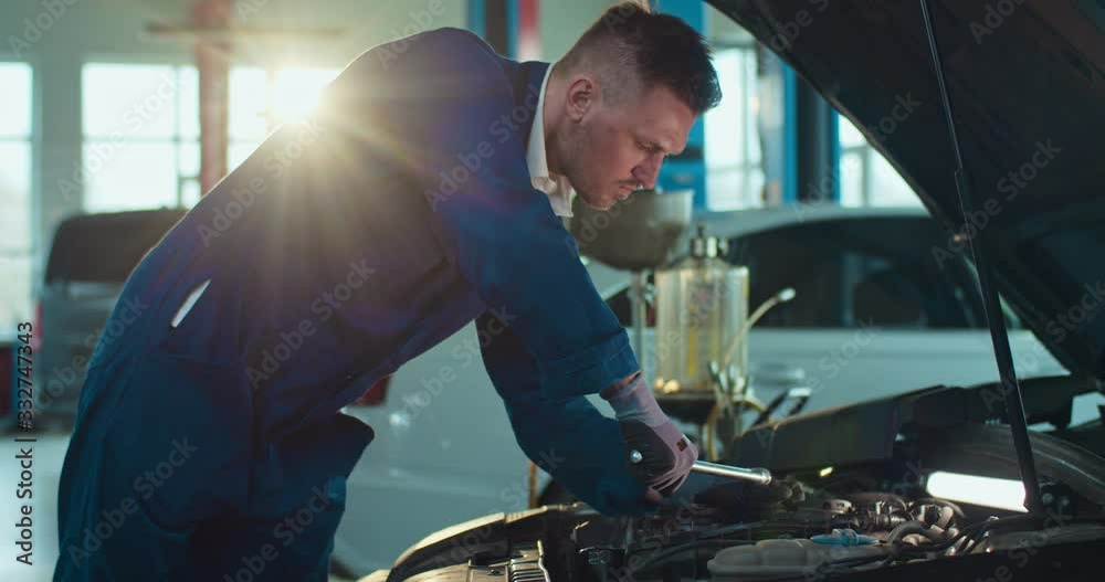 Male Caucasian young auto mechanic standing at open car and repairing breaks in big garage. Man in uniform and goggles working in automobile repairment salon indoor. Transport service concept. Male Caucasian young auto mechanic standing at open car and repairing breaks in big garage. Man in uniform and goggles working in automobile repairment salon indoor. Transport service concept.