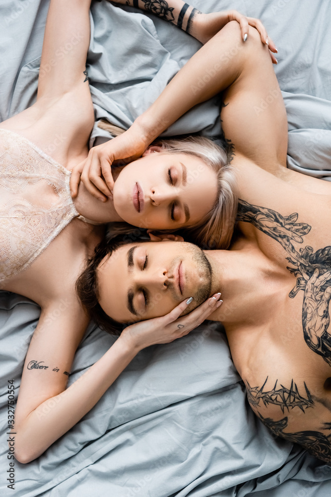 Top view of tattooed couple with closed eyes touching each other on bed