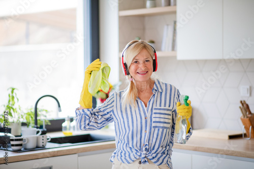 Portrait of senior woman with headphones and gloves cleaning indoors at home.