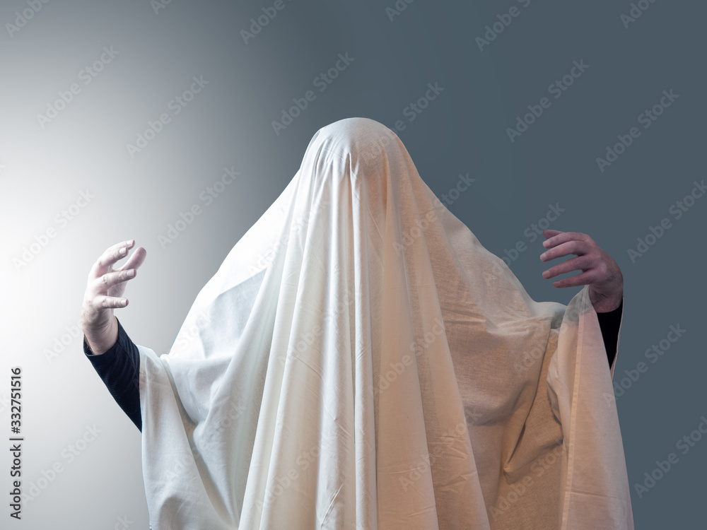 The man, covered in white drapery like a ghost, pulled his hands out from under it against a gray background with light on the left