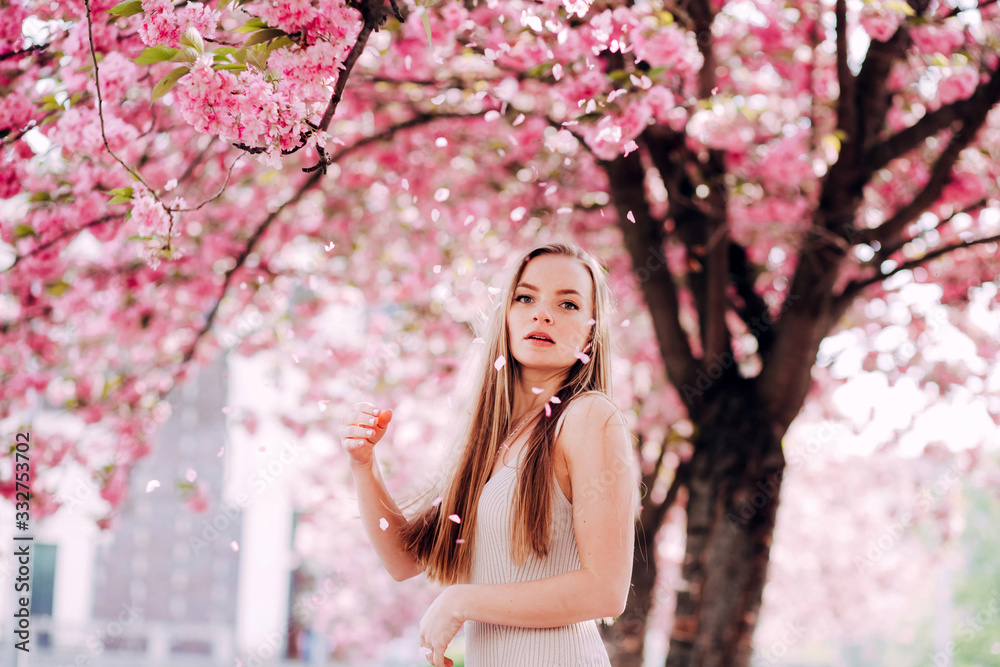 Closeup portrait of a lovely girl in a park with blooming japanese sakura trees. Romantic young blonde in a dress posing on a background of spring flowering trees. Flying cherry petals.