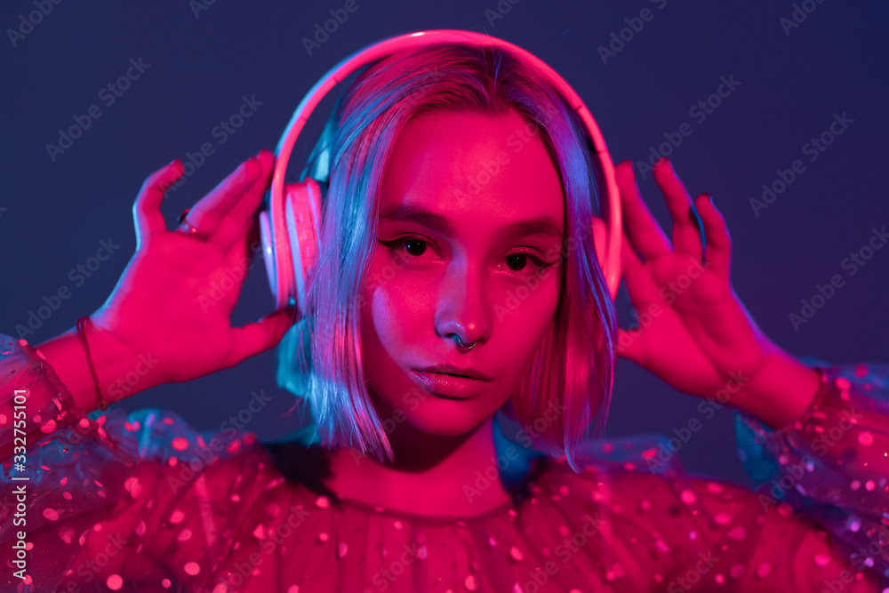 Gorgeous lady with dyed hair listening music in headphones and singing with neon light background. Charming hipster girl dancing with eyes closed.