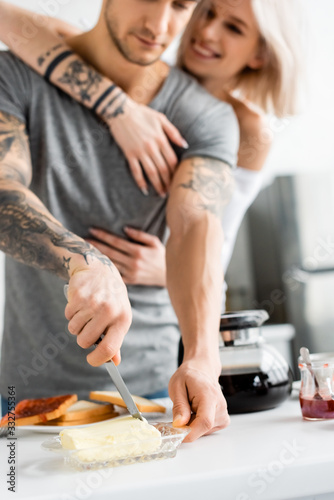 Selective focus of tattooed man cutting butter near smiling girlfriend in kitchen